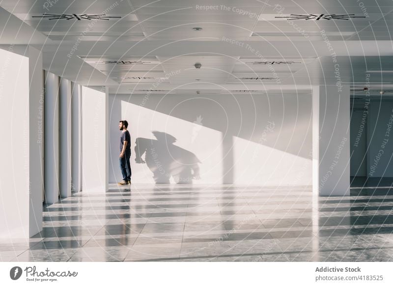 Man standing near window in spacious office hall man empty facility shadow sunlight geometry male interior shape hallway white wall modern design minimal line