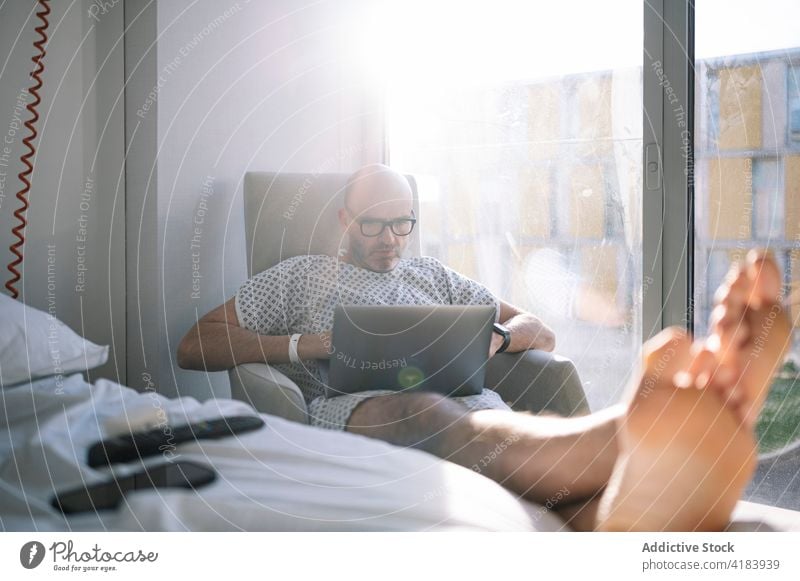 Focused male patient working on laptop on chair in ward man armchair concentrate workaholic hospital medicine device clinic watch professional digital sick ill