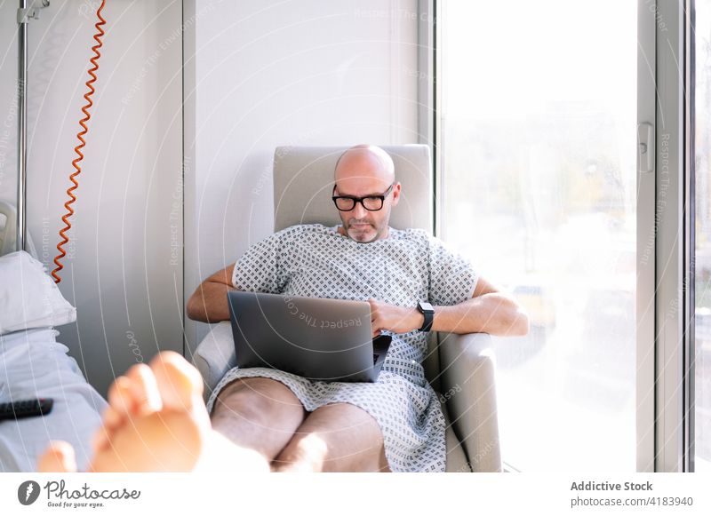 Focused male patient working on laptop on chair in ward man armchair concentrate workaholic hospital medicine device clinic watch professional digital sick ill