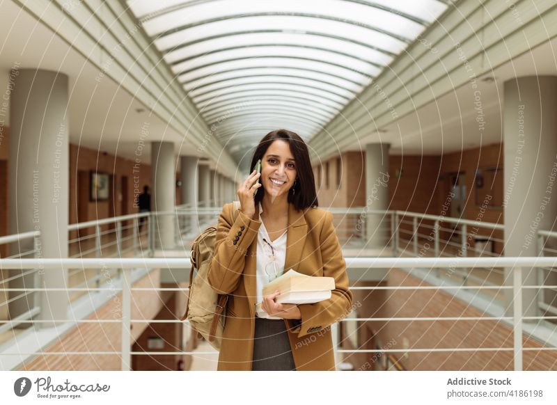 Happy woman talking on smartphone in hall of university building student hallway pile book speak using female ethnic mobile education conversation corridor call