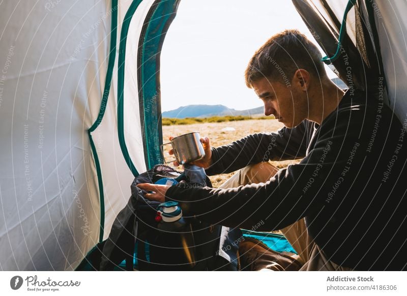 Young guy drinking hot beverage in camping tent man hot drink hike travel rest calm camper nature mountain hill trekking male young casual thermos tourism