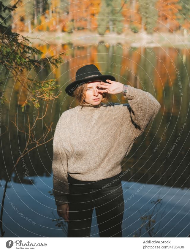 Stylish traveler in hat on shore against lake tourist cool style trip reflection tree woman sky stylish apparel garment confident trendy water mirror beautiful