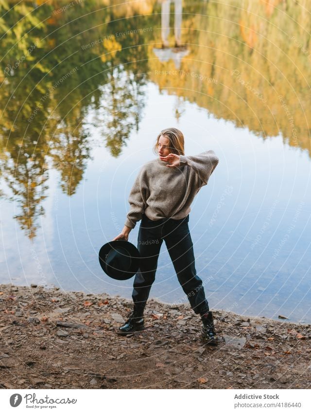 Stylish traveler in hat on shore against lake tourist cool style trip reflection tree woman sky stylish apparel garment confident trendy water mirror beautiful