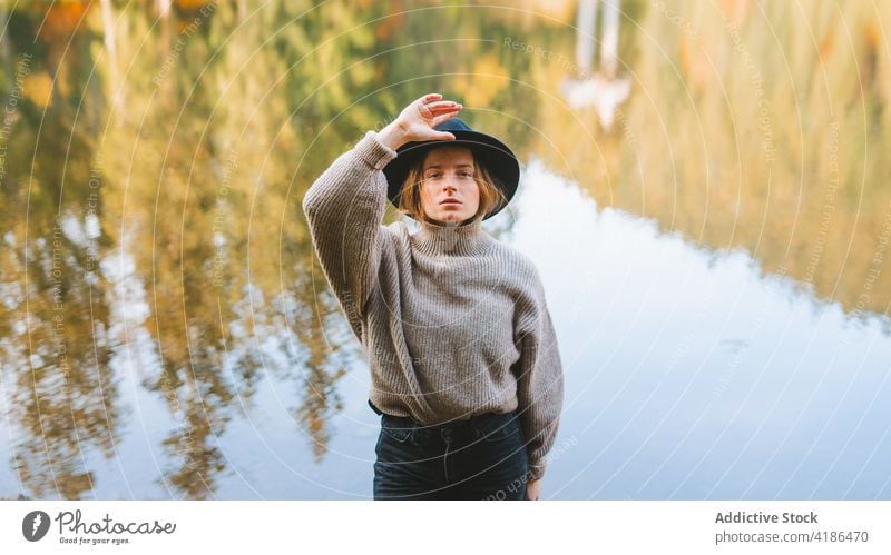 Stylish traveler in hat on shore against lake tourist cool style trip reflection tree woman sky stylish apparel garment confident trendy water mirror beautiful