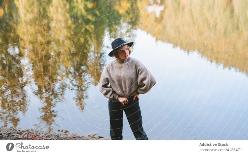 Stylish traveler in hat on shore against lake tourist cool style trip reflection tree woman sky stylish apparel garment confident trendy water mirror beautiful