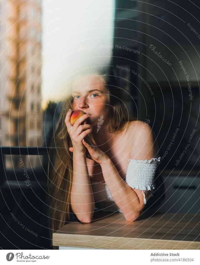 Beautiful woman with peach standing in modern kitchen dreamy snack serene positive lean on peaceful feminine counter tranquil charming bare shoulders attractive