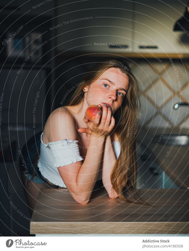 Beautiful woman with peach standing in modern kitchen dreamy snack serene positive lean on peaceful feminine counter tranquil charming bare shoulders attractive