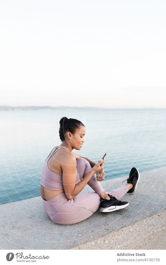 Ethnic sportswoman surfing internet on smartphone against sea on embankment online break nature legs crossed using gadget sky device ethnic rest sneakers