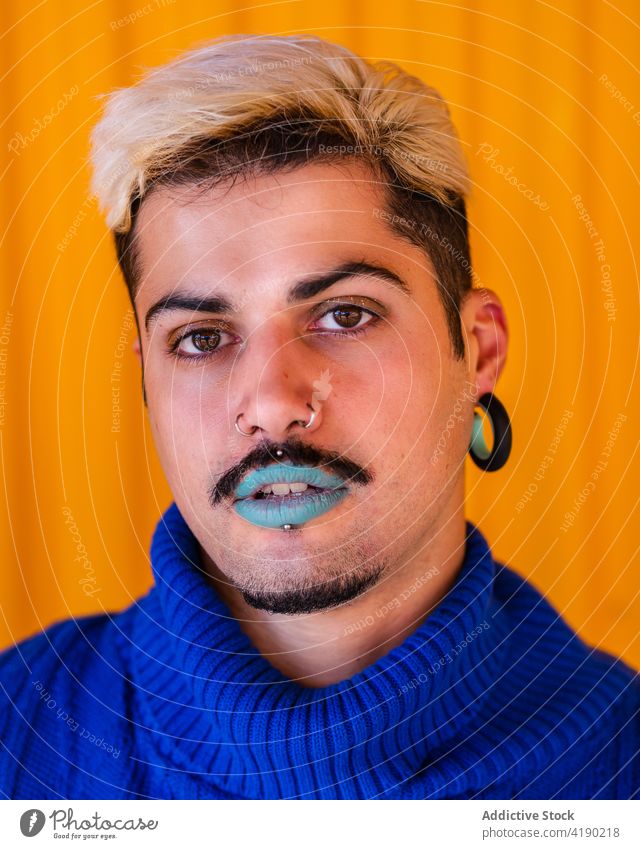 Stylish man with makeup looking at camera androgynous trendy model style fashion alternative apparel visage male blue lip lipstick emotionless outfit yellow