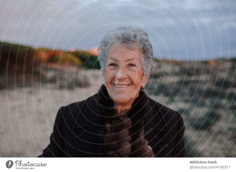 Positive senior woman smiling while resting on sandy seashore in evening smile relax beach tourist happy portrait holiday glad coast cloudy sky female elderly