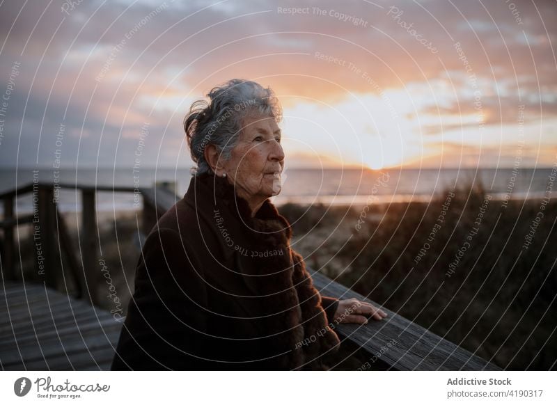 Senior lady admiring sunset over ocean from boardwalk woman admire seascape beach traveler enjoy nature vacation relax female elderly casual wooden pier