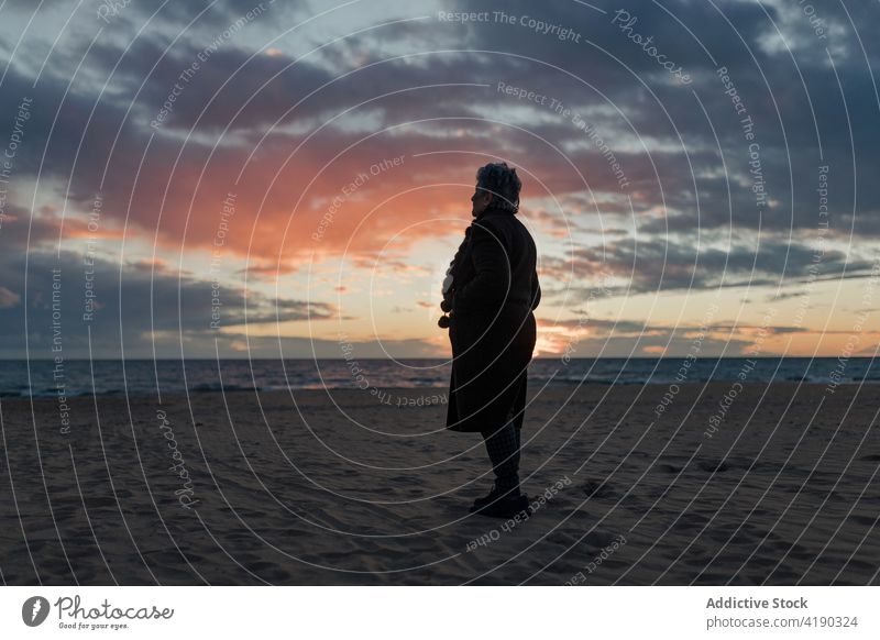 Anonymous calm elderly woman recreating on sandy seashore at sundown admire sunset beach enjoy relax alone nature landscape seascape female senior warm clothes