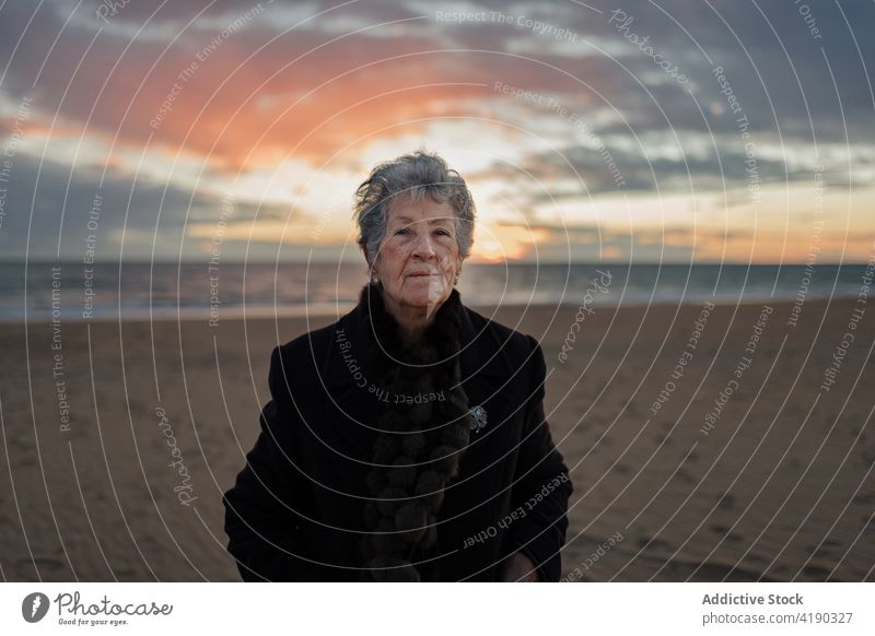 Calm elderly woman recreating on sandy seashore at sundown admire sunset beach calm enjoy relax alone nature landscape seascape senior warm clothes casual coast