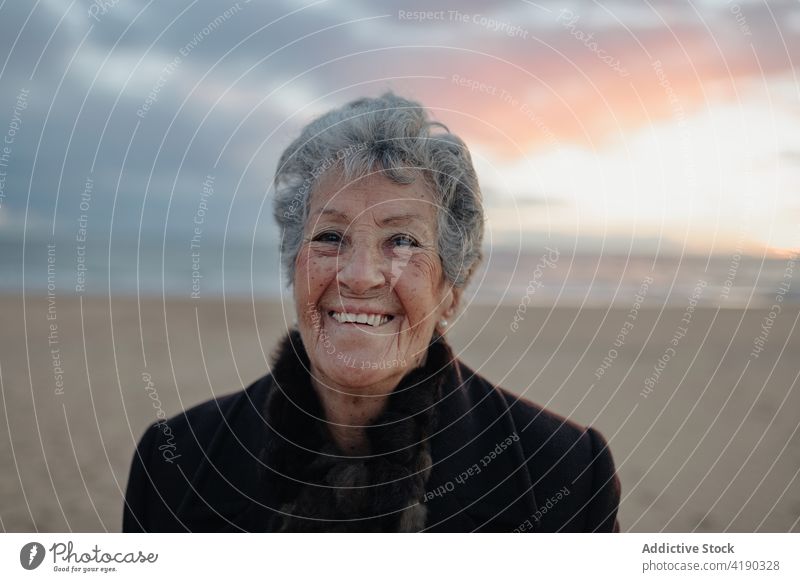 Calm elderly woman recreating on sandy seashore at sundown admire sunset beach calm enjoy relax alone nature landscape seascape senior warm clothes casual coast