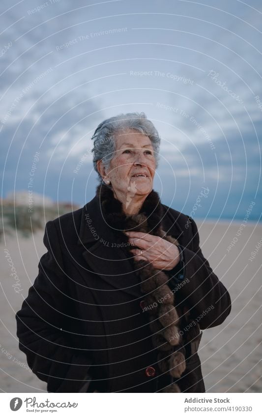 Senior woman resting on sandy seashore in evening relax beach tourist portrait holiday coast cloudy sky female elderly gray hair warm clothes casual ocean