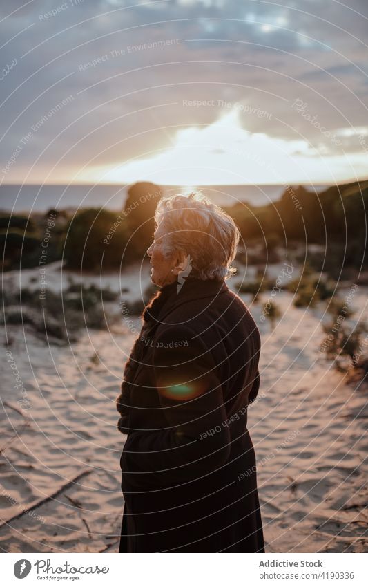 Senior woman resting on sandy seashore in evening relax beach tourist portrait holiday coast cloudy sky female elderly gray hair warm clothes casual ocean