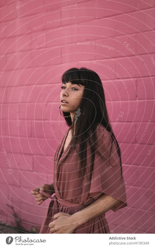 Fashionable young ethnic lady standing near pink wall and looking away woman pensive thoughtful style cool fashion calm street dreamy gorgeous brick wall