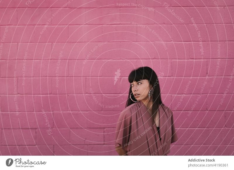 Fashionable young ethnic lady standing near pink wall and looking away woman pensive thoughtful style cool fashion calm street dreamy gorgeous brick wall