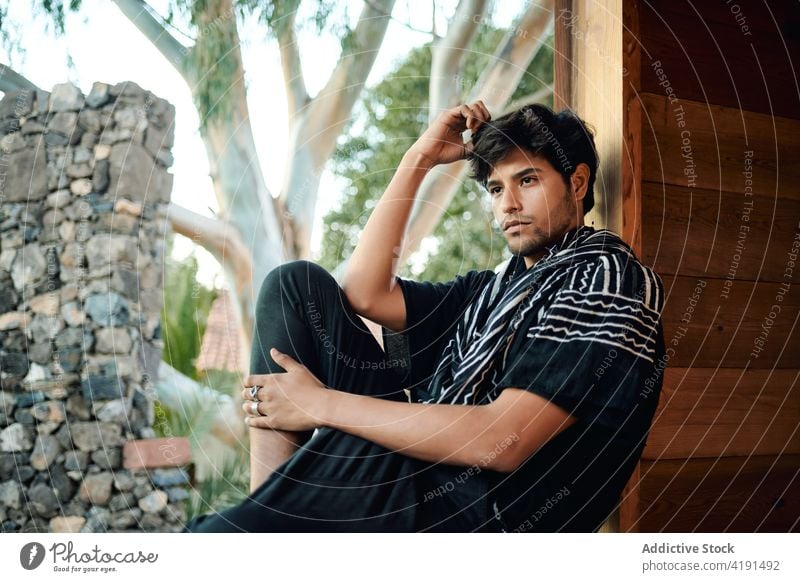 Hipster man in summer clothes sitting on windows sill surrounded by green vegetate fashion style barefoot model garment confident outfit wear young ethnic male