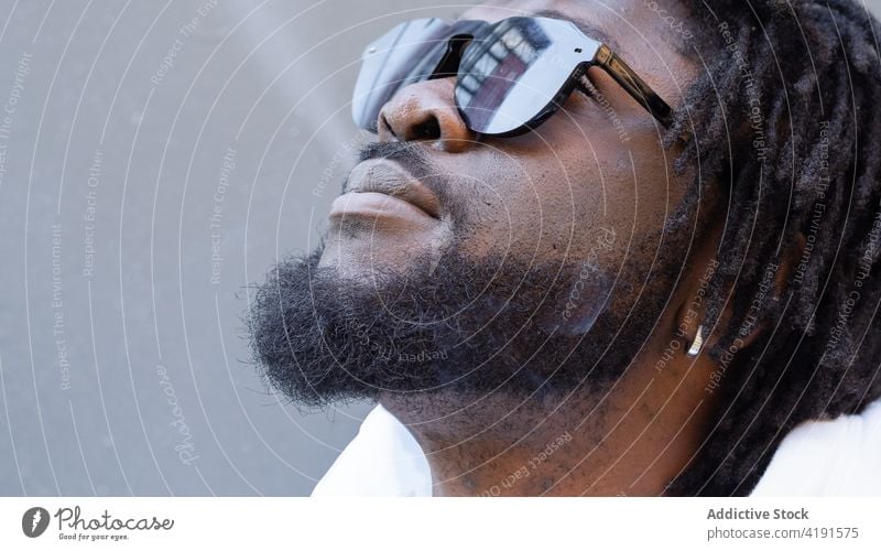 Crop dreamy black hipster man in sunglasses on urban road masculine brutal macho reflection street portrait earring mustache beard cool building town house