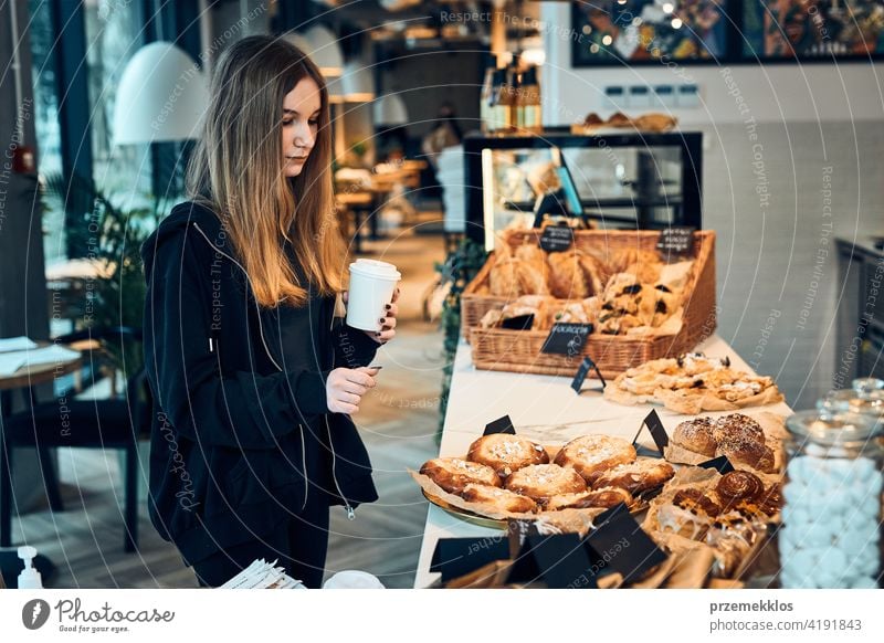 Woman holding cup with coffee looking at pastry, buns, cakes and cookies and waiting for the order. Girl buying a sweet food and hot drink to go. Young woman having a break doing shopping in a coffee shop