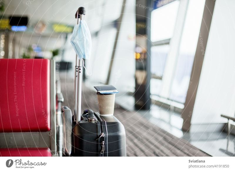 Next to the seats in an empty airport is a suitcase, on it is a glass of coffee, hanging a medical protective mask. Coronavirus, social distance, isolation