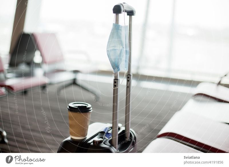 Next to the seats in an empty airport is a suitcase, on it is a glass of coffee, hanging a medical protective mask. Coronavirus, social distance, isolation