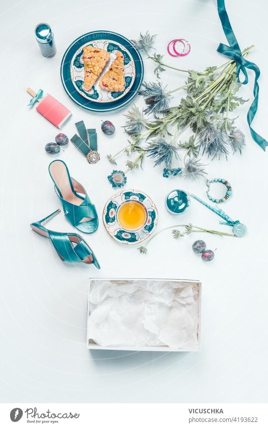 Female lifestyle flat lay with blue new high heels shoes, bouquet of flowers, a cup of tea, cakes, jewelry, cosmetics, empty box, fruits on white desk . Top view. Beauty and fashion concept