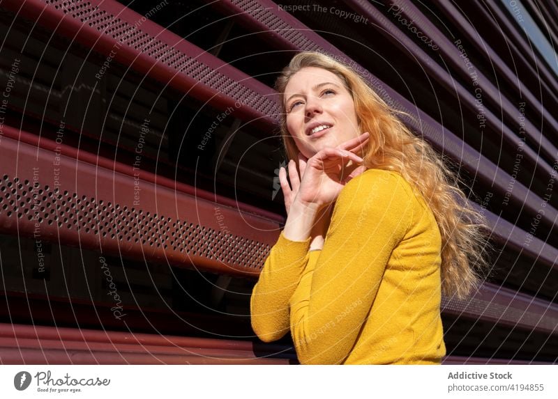 Contemplative young woman standing near metal fence on city street pensive thoughtful blonde calm appearance style sunlight facade female casual long hair