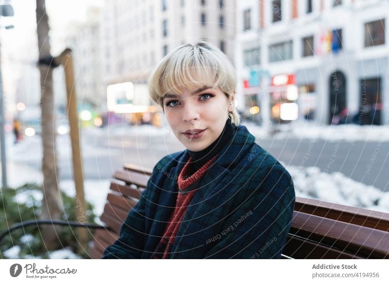 Positive young woman resting on bench in modern city street warm clothes season autumn urban style beautiful trendy casual pretty contemporary coat lifestyle