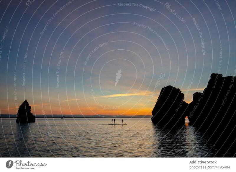 Unrecognizable sportspeople on paddle board near rocks in ocean sup board sea sky cloudy nature seascape silhouette highland landscape evening sunset ripple