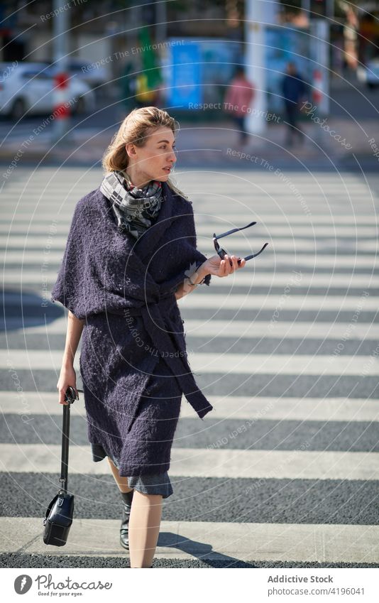 Stylish woman crossing urban road stylish flying hair stroll fashion town lifestyle sunglasses blonde crosswalk black cloth accessory hold individuality modern