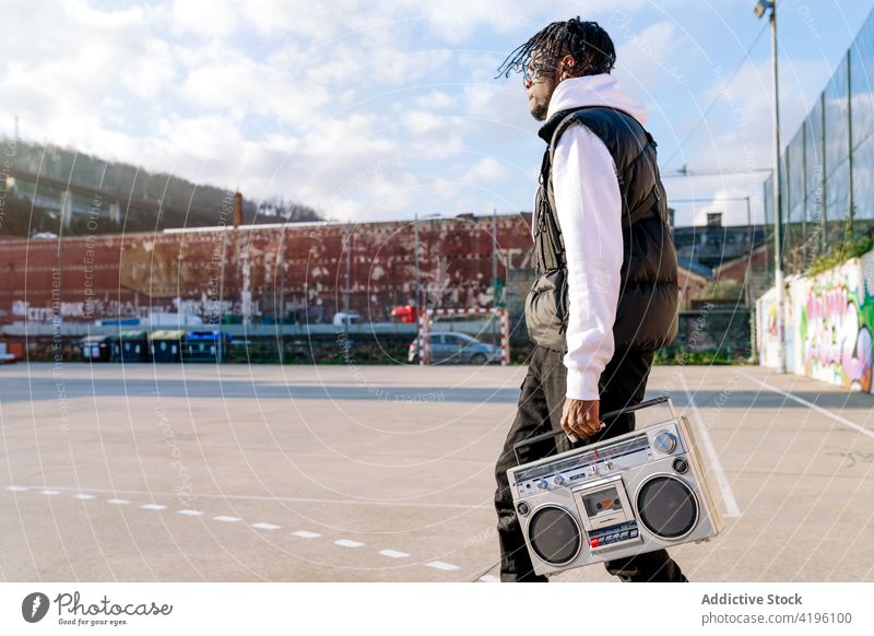 Black hipster man carrying retro cassette recorder on urban pavement style hip hop subculture walk aged trendy apparel vintage town black african american