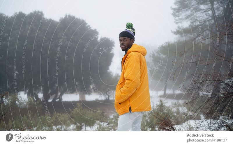 Serious male standing among trees in winter forest man snow style nature park cold serious flora haze outerwear pensive hand in pocket warm clothes environment