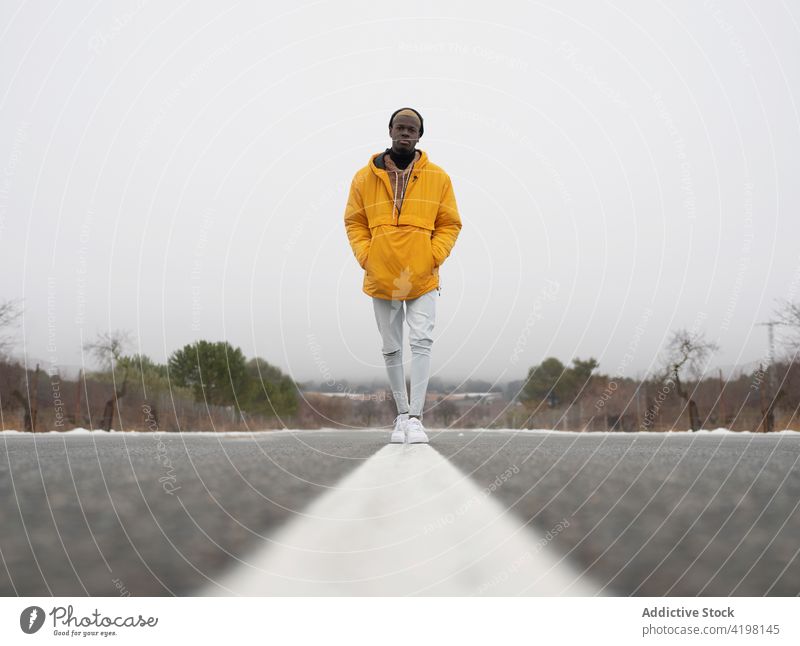 Stylish black man standing on road in nature style confident environment snow cold individuality cool male roadway leafless hand in pocket tree roadside route