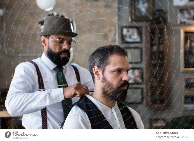 Ethnic stylist with hipster client preparing for procedure in barbershop hairstylist prepare profession masculine brutal virile stylish men formal apparel