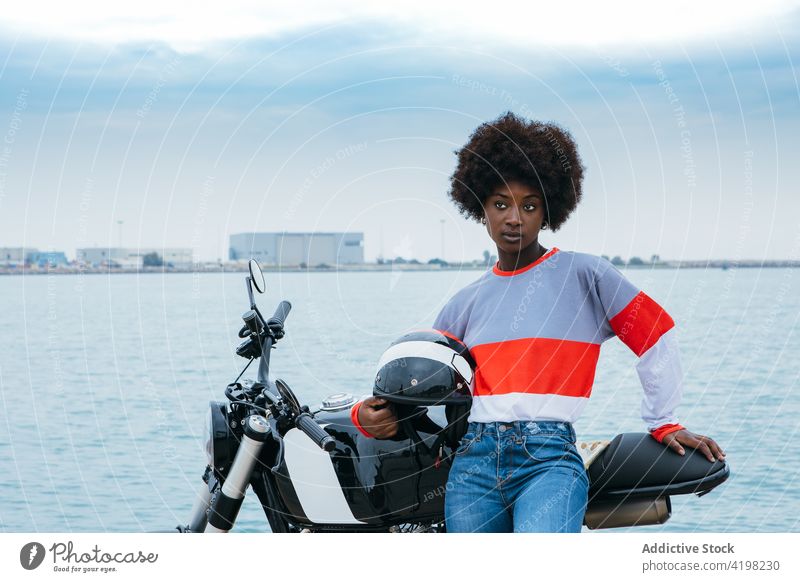 Serious young ethnic woman wearing helmet while sitting on motorbike motorcycle seaside appearance personality trendy embankment alone biker cool travel vehicle