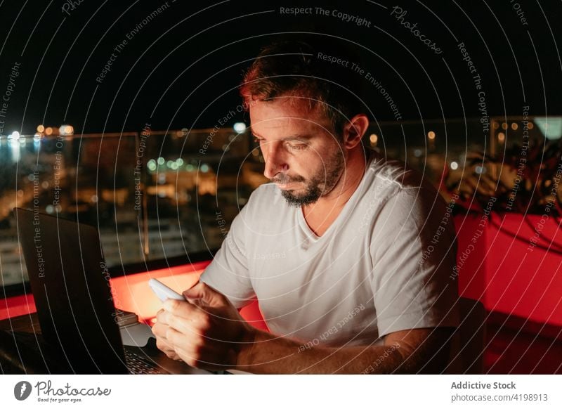 Focused man using smartphone and laptop during online work on rooftop at night message remote evening freelance concentrate communicate connection male young