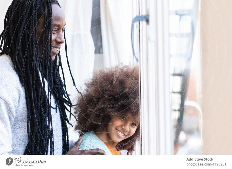 Black father with daughter standing near window close cheerful carefree bonding positive tender smile hug girl man dad childhood kid afro parent casual love