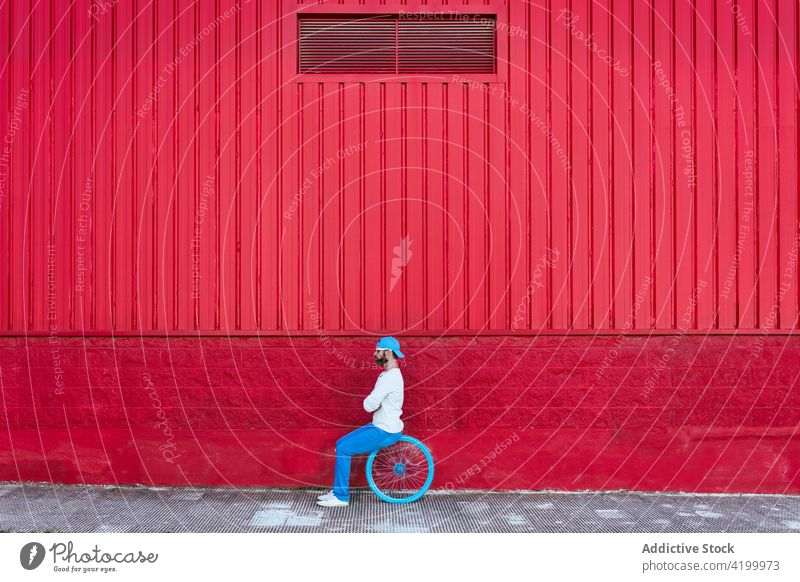 Hipster man sitting in wheel on city street building male beard hat hipster urban modern hobby lifestyle bicycle outdoors masculine sunglasses red background