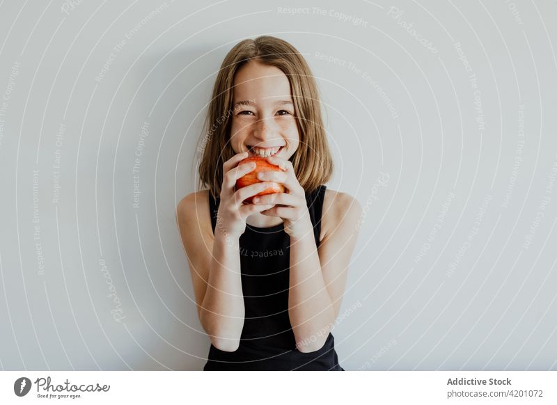 Delighted kid biting apple against white wall girl bite cheerful smile eat fresh fruit vitamin enjoy portrait meal happy ripe child preteen casual top