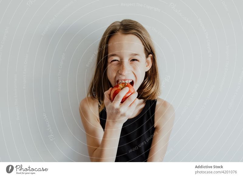 Delighted kid biting apple against white wall girl bite cheerful smile eat fresh fruit vitamin enjoy portrait meal happy ripe child preteen casual top