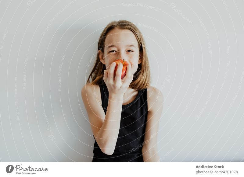 Delighted kid biting apple against white wall girl bite cheerful smile eat fresh fruit vitamin enjoy portrait meal happy ripe child preteen casual top