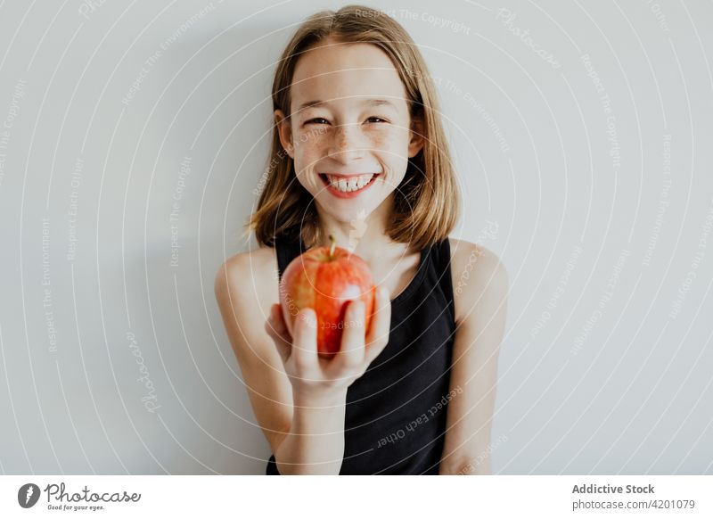 Delighted kid biting apple against white wall girl bite cheerful smile eat fresh fruit vitamin enjoy portrait meal happy ripe child preteen casual top