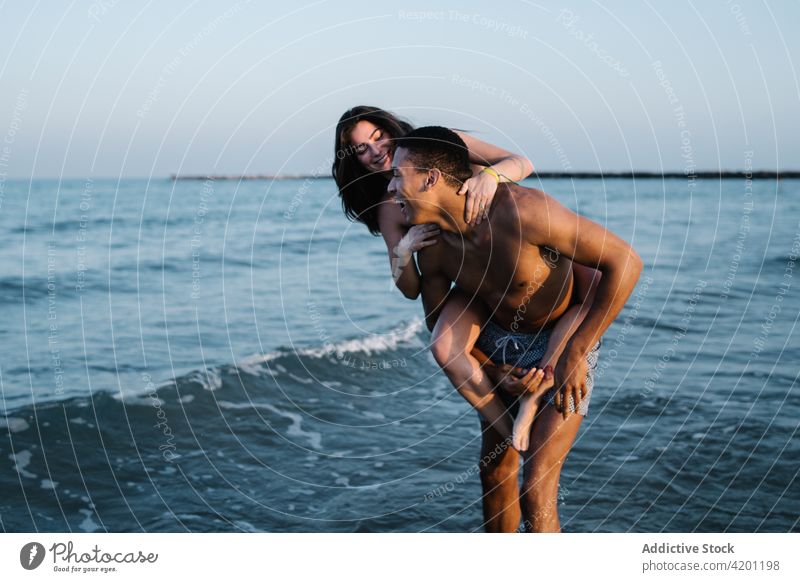 Cheerful black man giving girlfriend piggyback ride against sea couple relationship having fun honeymoon ocean trip vacation happy wavy boyfriend love smile