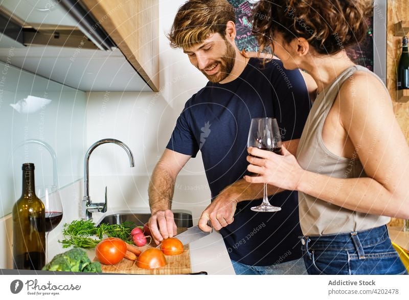 Couple in kitchen with wineglass while cutting vegetables couple cook tomato drink friend counter lifestyle young cabinet home red stove knife sink tap alcohol