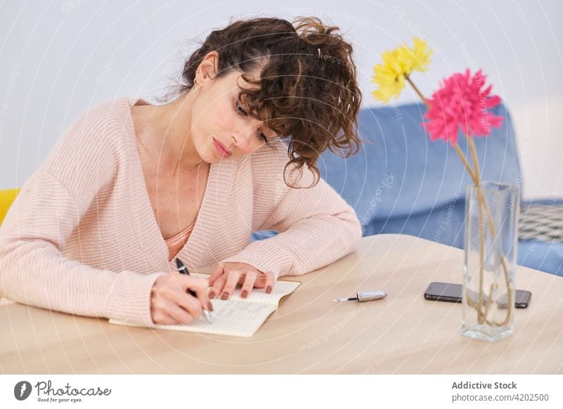 Thoughtful woman writing notes in copybook at home write notepad room take note smartphone vase flower thoughtful young female table casual notebook pen