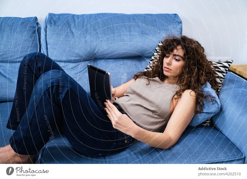 Young woman using tablet while having rest on couch lying browsing sofa room surfing internet relax free time female casual living room chill home comfort