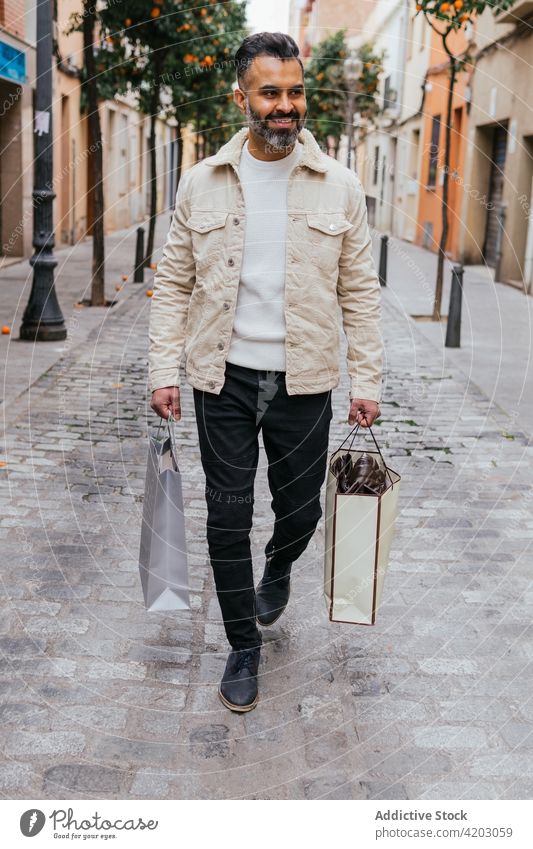 Smiling Hispanic shopper with gift bags walking on city street shopping bag smile masculine style man urban lifestyle hispanic ethnic glad enjoy present modern