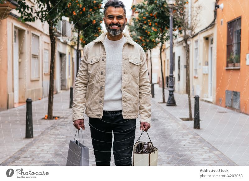 Smiling Hispanic shopper with gift bags walking on city street shopping bag smile masculine style man urban lifestyle hispanic ethnic glad enjoy present modern